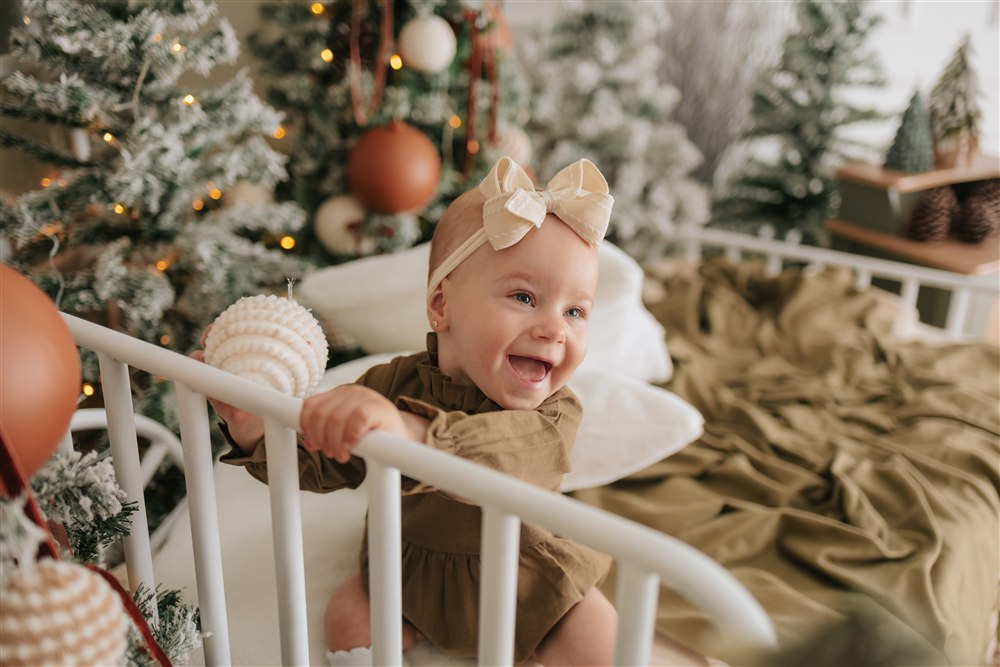 Bebê sorrindo entre árvores de Natal, luzes quentes e enfeites